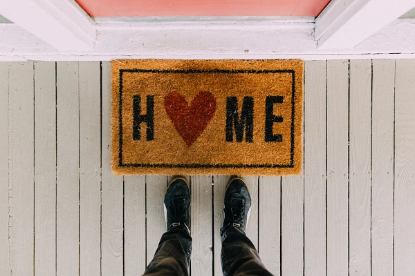 A person stands on a doormat that spells "HOME" with a heart symbol replacing the letter "O."0